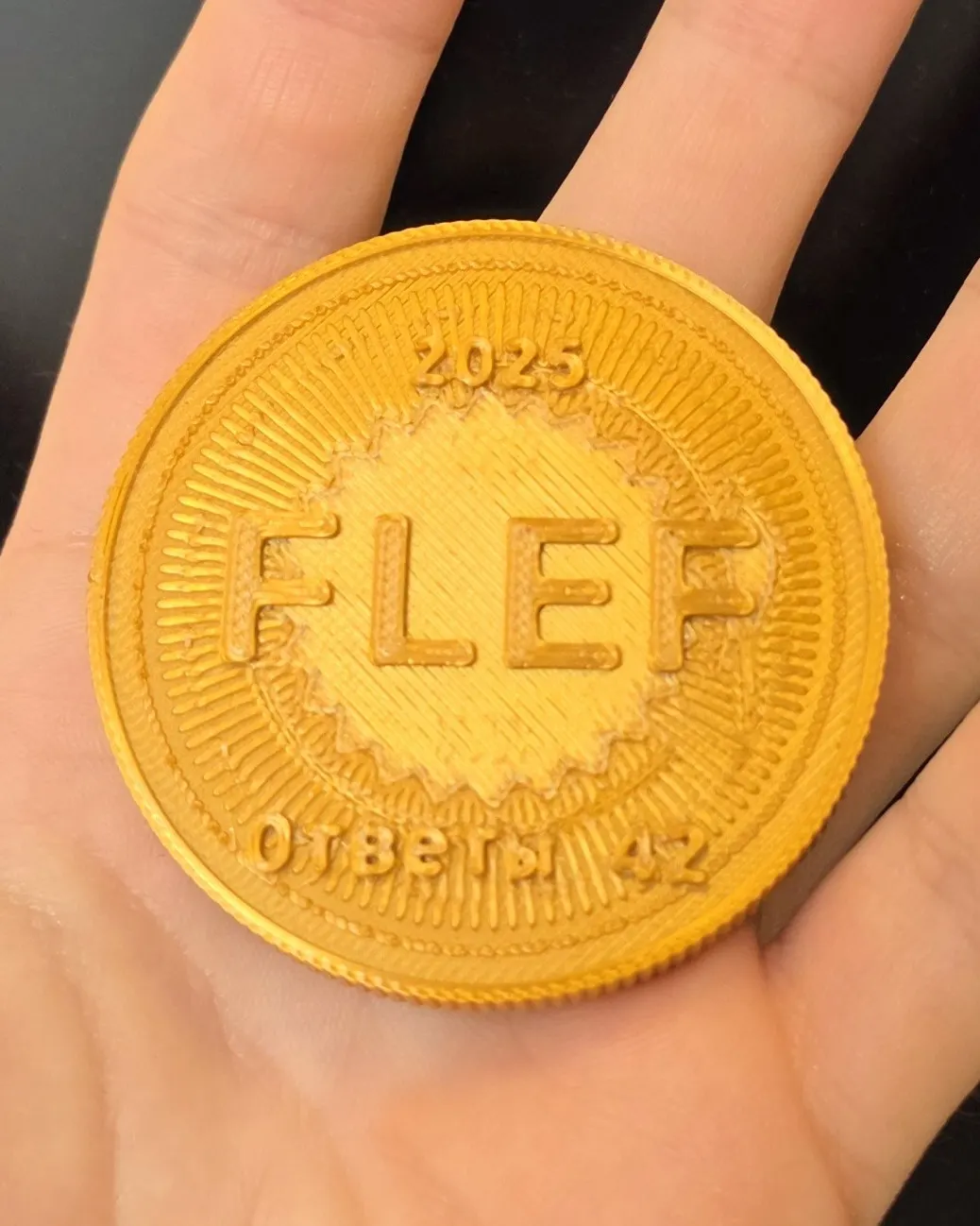 Zlato rumen natisnjen kovanec z napisom FLEF, v dlani